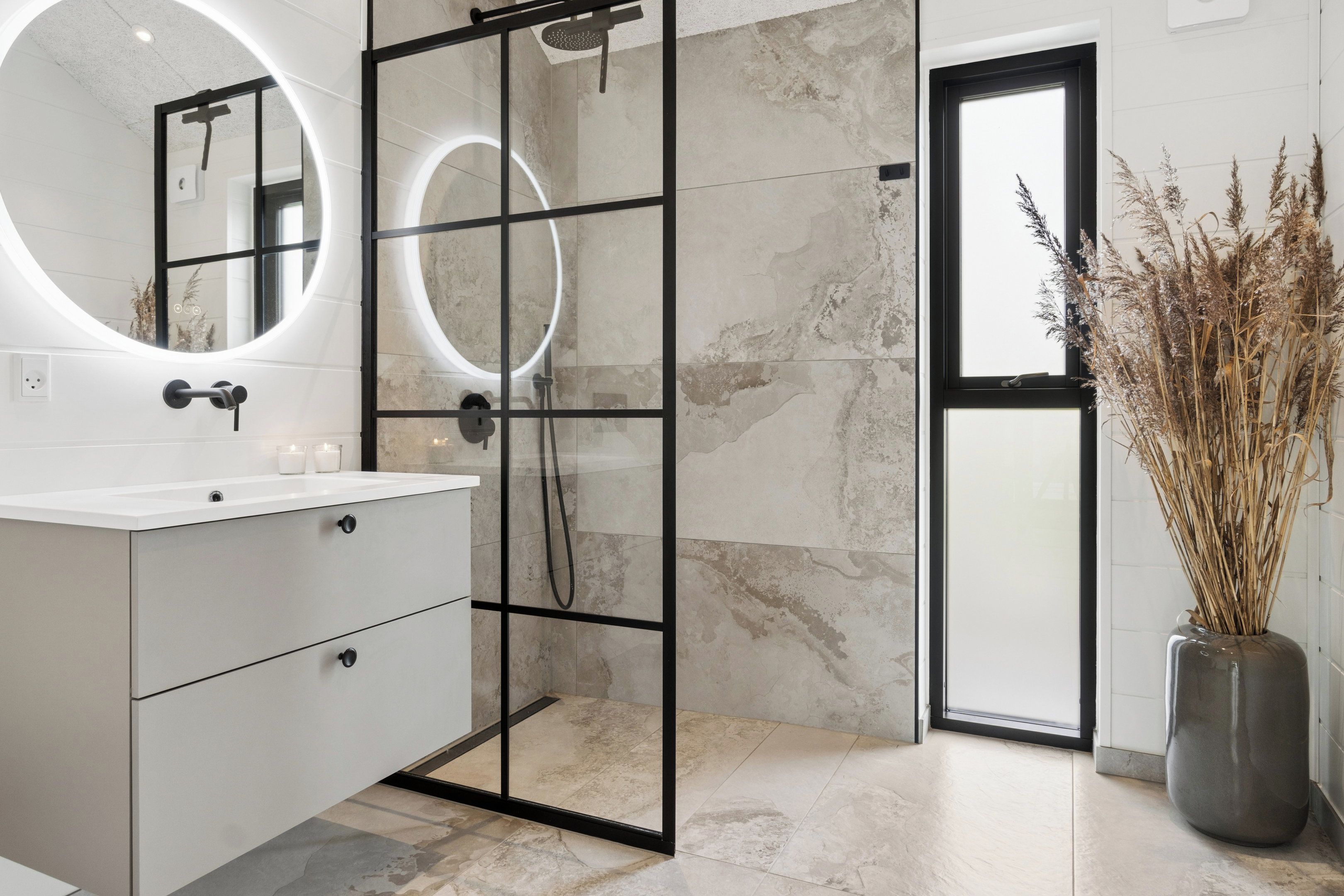 Une salle de bain moderne avec une douche à parois de verre, un grand miroir et un vase contenant des plantes séchées.