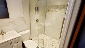 Ein Badezimmer mit einer gläsernen Duschkabine, einer weißen Toilette und einem Waschtischbereich mit Marmorfliesen.