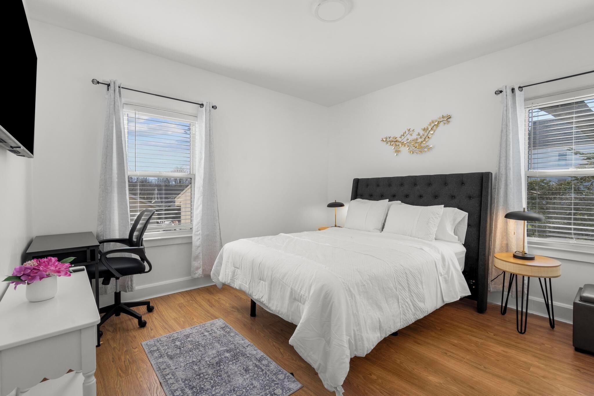 Een slaapkamer met een groot bed, een bureau met een stoel, een klein tafeltje met een lamp, en een decoratief wandkunstwerk.