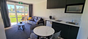 A modern room with a sofa, a small table, a kitchen area with a microwave, and a view of a green outdoor area. - Deluxe Family Apartment with Spa Bath & Alpacas (Rotorua)