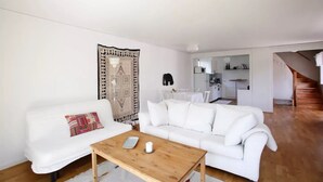 Una sala de estar con un sofá blanco, una mesa de centro de madera y un tapete estampado. Se ve una cocina al fondo con armarios blancos y un comedor.