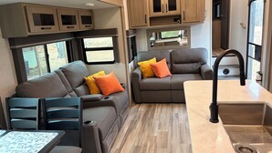 A compact RV interior with a kitchenette, two seating areas with cushions, and a dining table.