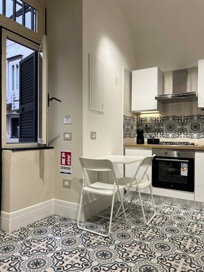 A compact kitchen with a window, a white table, two white chairs, a patterned backsplash, and a small oven. - Old town apartment, new and cozy (Napoli)
