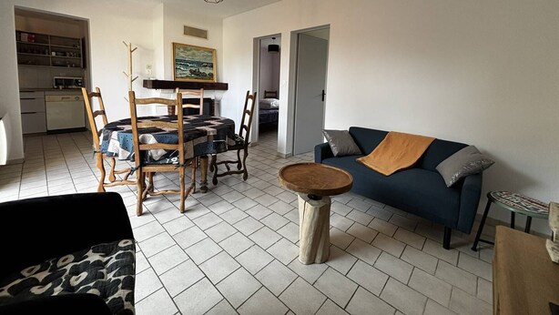 Een woonkamer met een eettafel, stoelen, een bank en een salontafel. Aan de muur hangt een schilderij en er is een keuken in de achtergrond.
