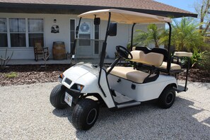 Une voiturette de golf est garée devant un bâtiment doté d’un porche, d’une chaise en bois et d’un tonneau.