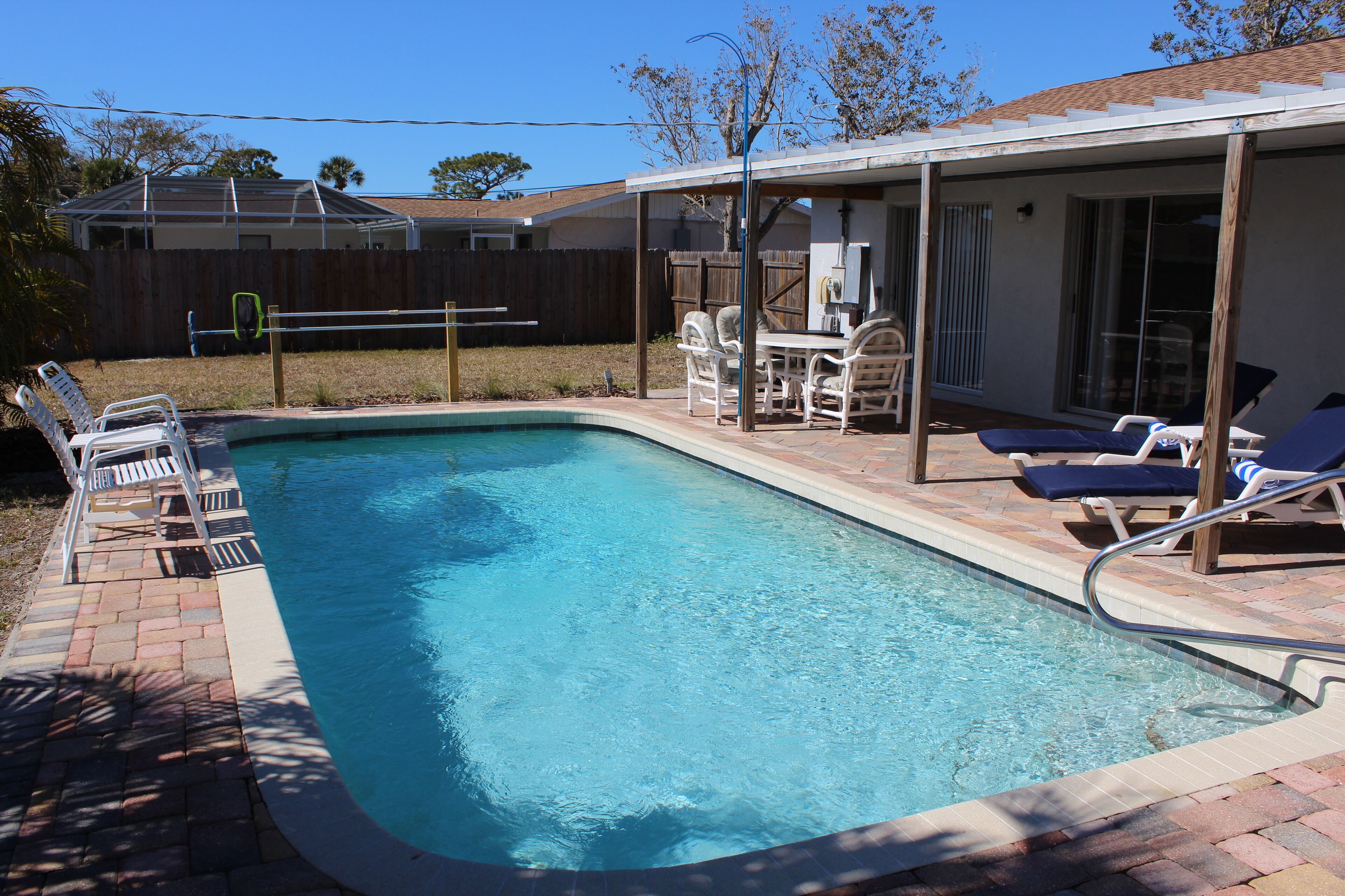 Une piscine rectangulaire bordée de carreaux, entourée d’un patio couvert aménagé avec du mobilier d’extérieur et une clôture en bois en arrière-plan.