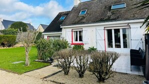 A two-story house with a sloped roof, white exterior walls, and red window shutters. There is a small garden with shrubs and a tree in front of the house. - Maison tout confort, proche plage et centre-ville (QUIBERON)