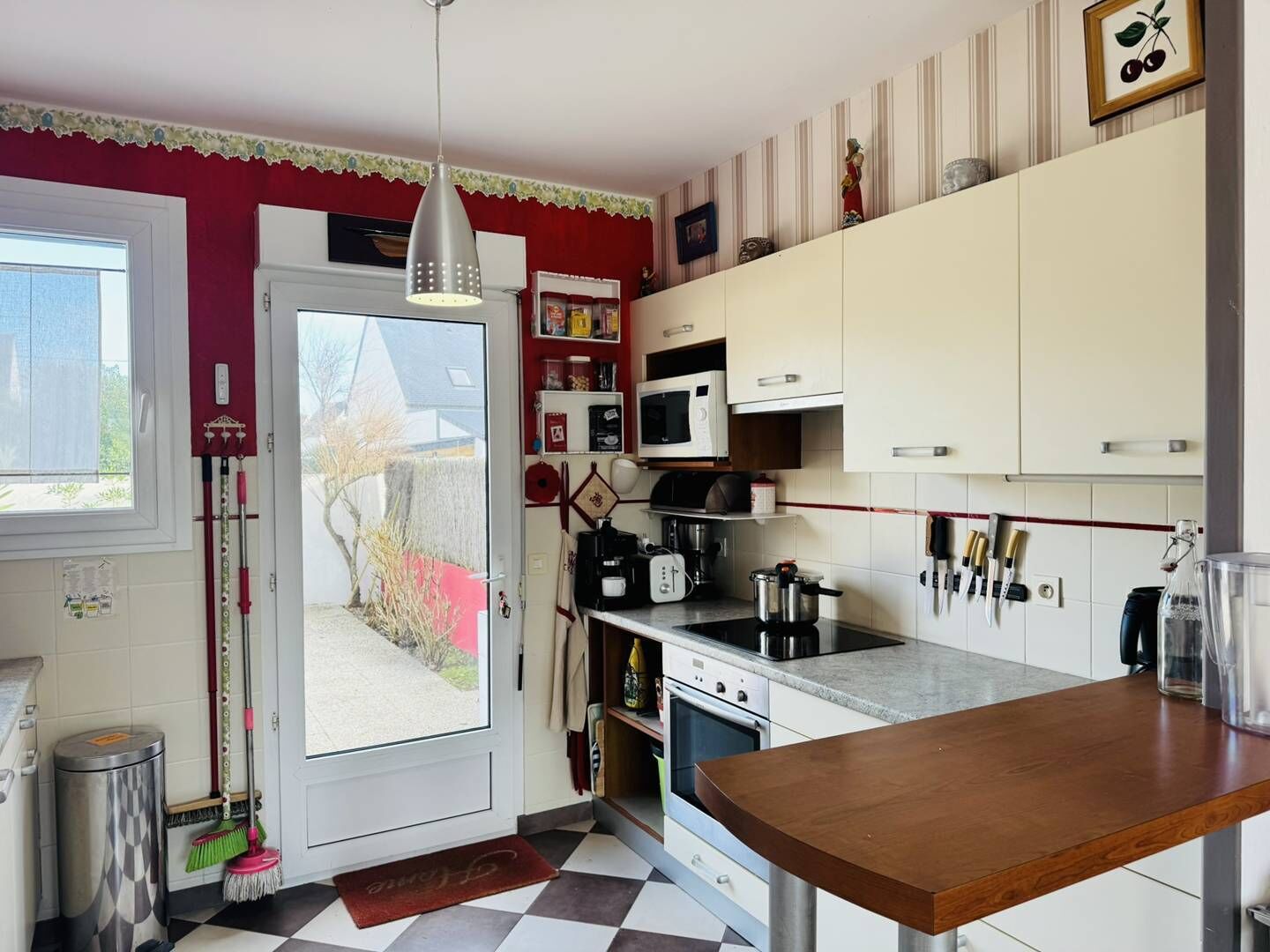 Une cuisine équipée d’un four à micro-ondes, d’une cuisinière et de divers ustensiles. Une fenêtre offre une vue sur une maison et un arbre.