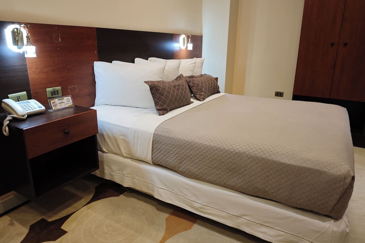 A neatly made bed with a quilted cover, a bedside telephone, a nightstand, and a wall-mounted light fixture.
