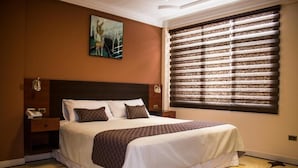 A neatly made bed with a headboard, a bedside table with a phone, and a window with blinds. - HOTEL PAIRUMANI (Cochabamba)
