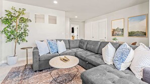 Een moderne woonkamer met een grijze modulaire bank, een ronde salontafel en een grote potplant.