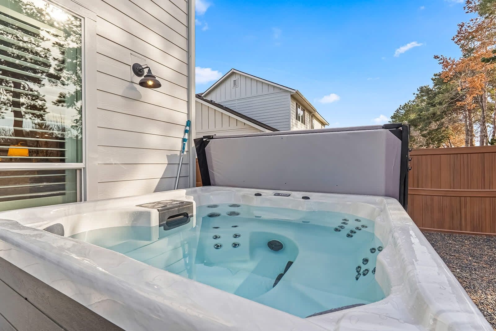 A hot tub with a control panel, a window with blinds, and a wooden fence in the background.