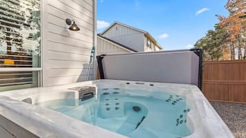 A hot tub with a control panel, a window with blinds, and a wooden fence in the background.