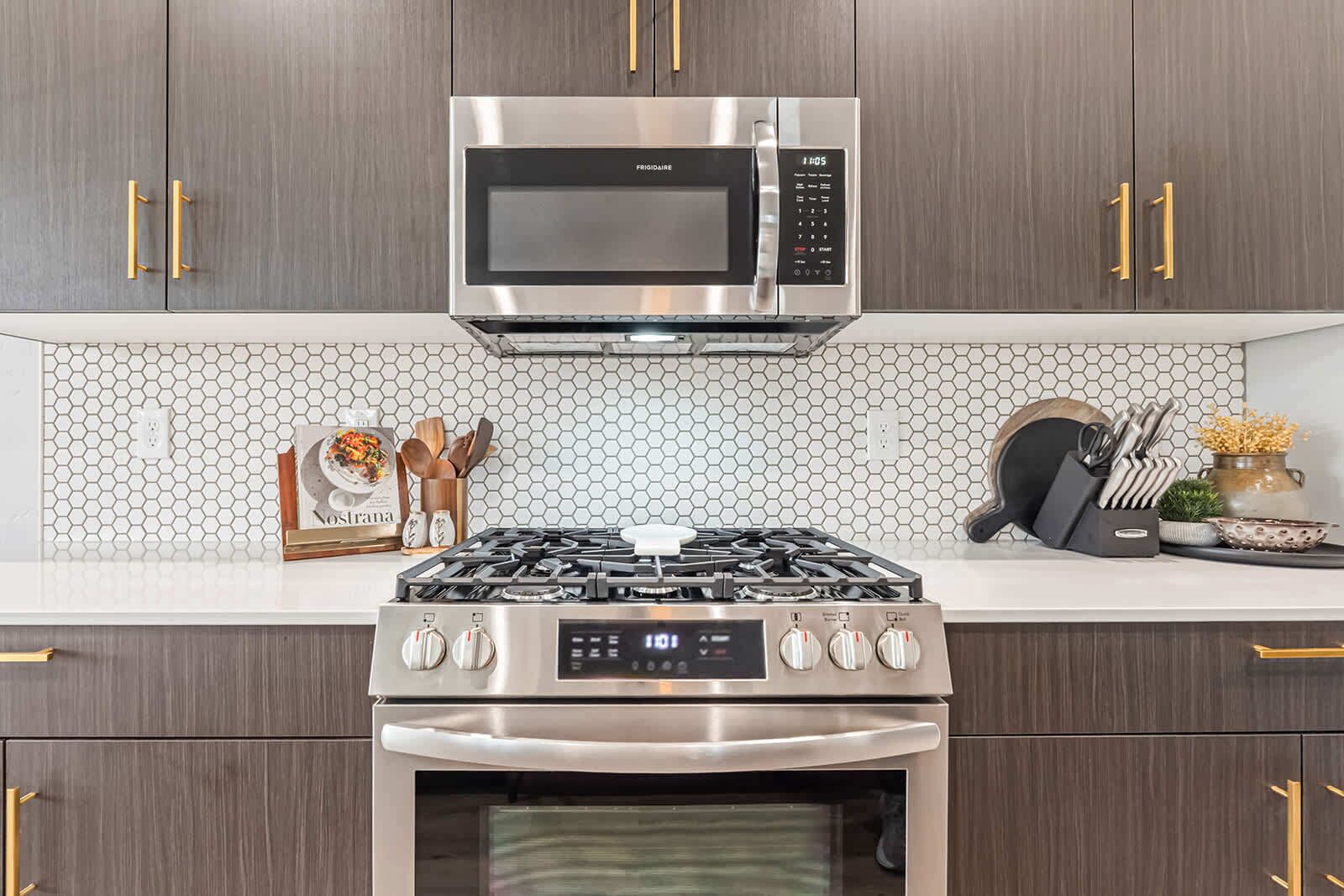 A kitchen with a stainless steel microwave, gas stove, and oven, wooden cabinets with gold handles, and a hexagonal tile backsplash.