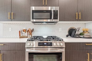 A kitchen with a stainless steel microwave, gas stove, and oven, wooden cabinets with gold handles, and a hexagonal tile backsplash. - Generous 4BR Home w Hot Tub & Game room (Boise)