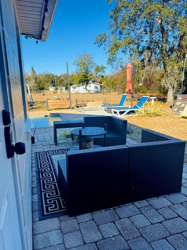 Un patio con un sofá de mimbre y una pequeña mesa, un espacio de estar techado al aire libre y vistas a un jardín vallado con árboles y una casa al fondo.