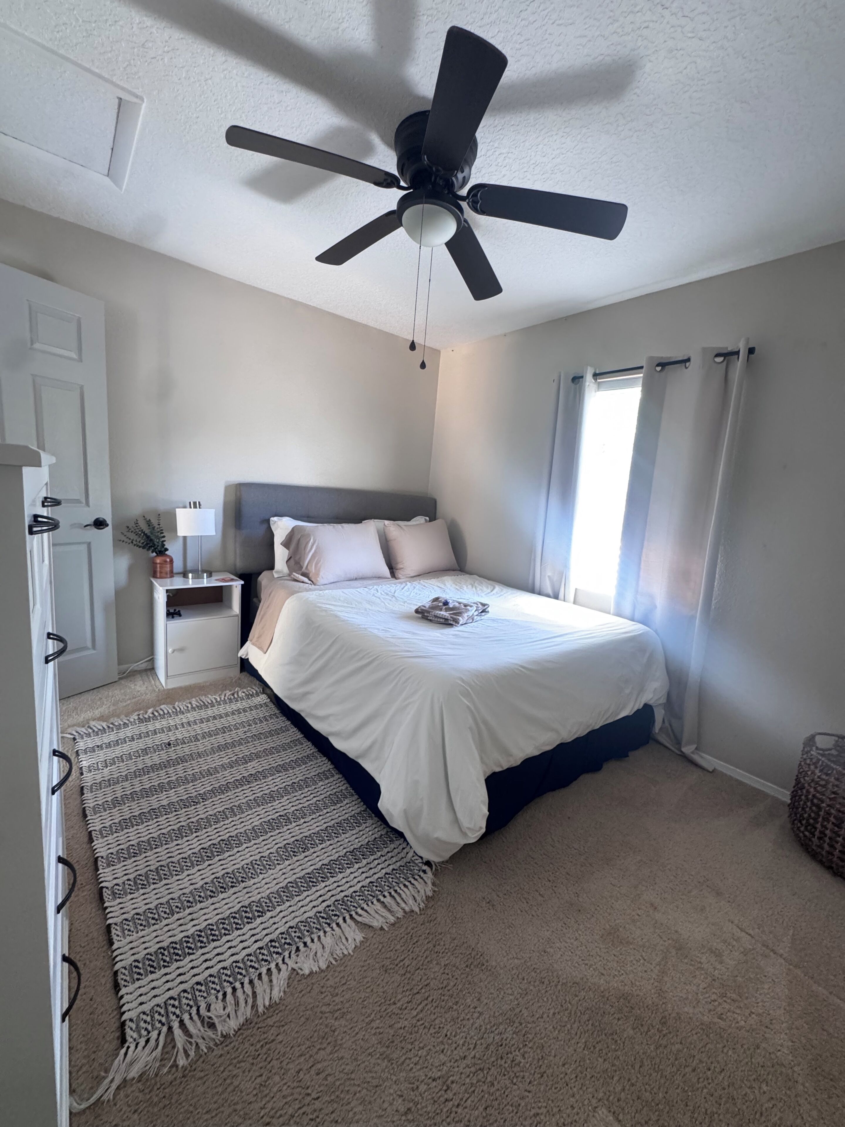 Un dormitorio con cama, ventilador de techo, mesita de noche, lámpara, ventana con cortinas y puerta.