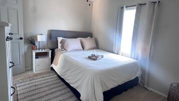 Un dormitorio con cama, ventilador de techo, mesita de noche, lámpara, ventana con cortinas y puerta.
