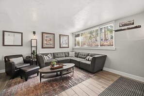 A living room with a sectional sofa, a coffee table, a side table, a rug, framed pictures, a lamp, and a window with blinds. - Park City Ski Condo Walk to Slopes and Main Street (Park City)