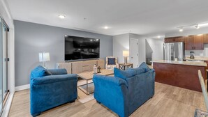 A modern living room with two blue sofas, a flat-screen TV, a wooden coffee table, and a kitchen area with a refrigerator and cabinets. - Serene Birmingham Retreat (Birmingham)