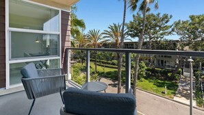 Ein Balkon mit Korbstuhl und kleinem Tisch, mit Blick auf einen gepflegten Garten mit Palmen und strahlend blauem Himmel.