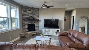 Una sala de estar con chimenea de piedra, un televisor de pantalla plana grande, un sofá de cuero marrón y un ventilador de techo.