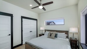 A bedroom with a bed, two bedside tables with lamps, a ceiling fan, and a window with a view of a house.