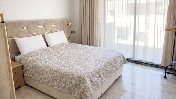 Una moderna camera d'albergo con un grande letto, un comodino in legno, un tavolino accanto al letto e un'ampia finestra con tende leggere.