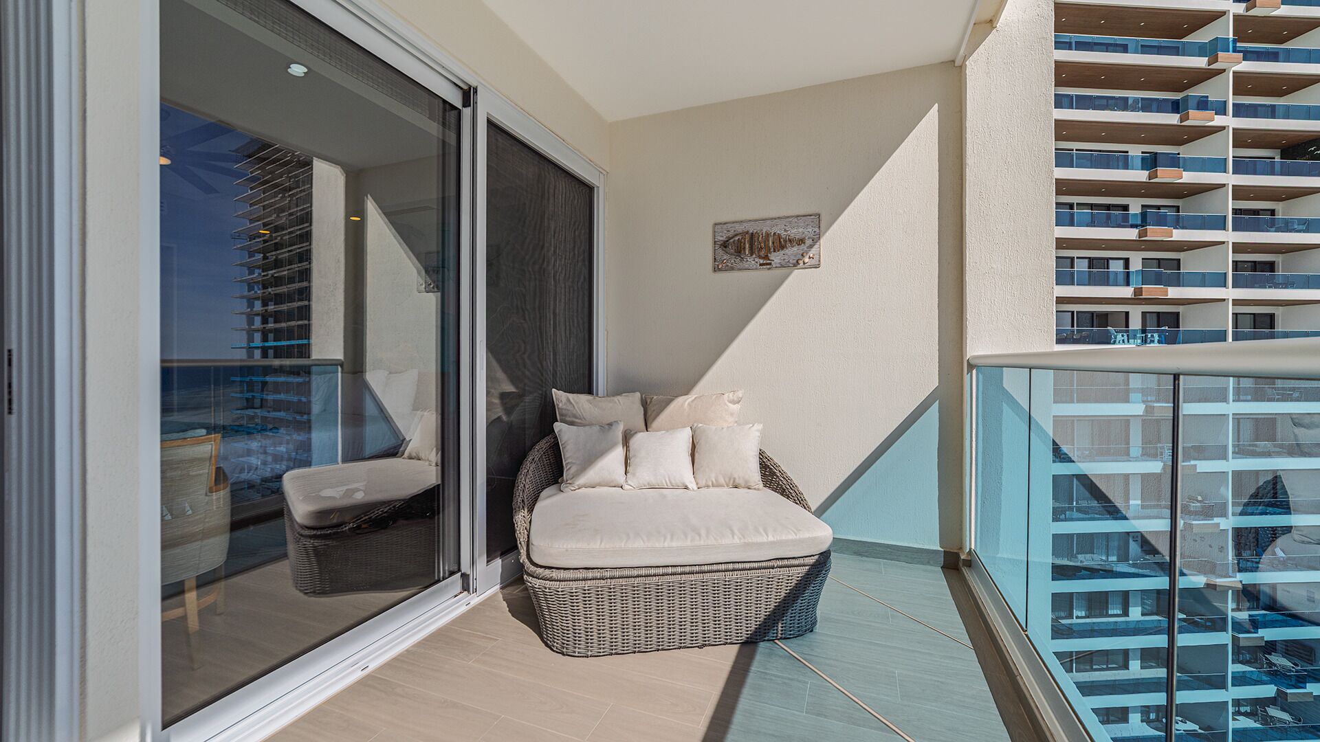 Ein Balkon mit einem Rattan-Sofa, Kissen, einer Glastür und Blick auf ein Gebäude mit Balkonen.
