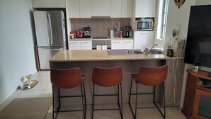 A compact kitchen with a refrigerator, microwave, and coffee maker. There are three stools and a TV on a wooden stand. - Water views luxury 2 bed fully furnished unit (Hope Island)