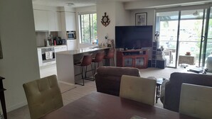 A modern living room with a dining area, kitchen, and a large TV. - Water views luxury 2 bed fully furnished unit (Hope Island)