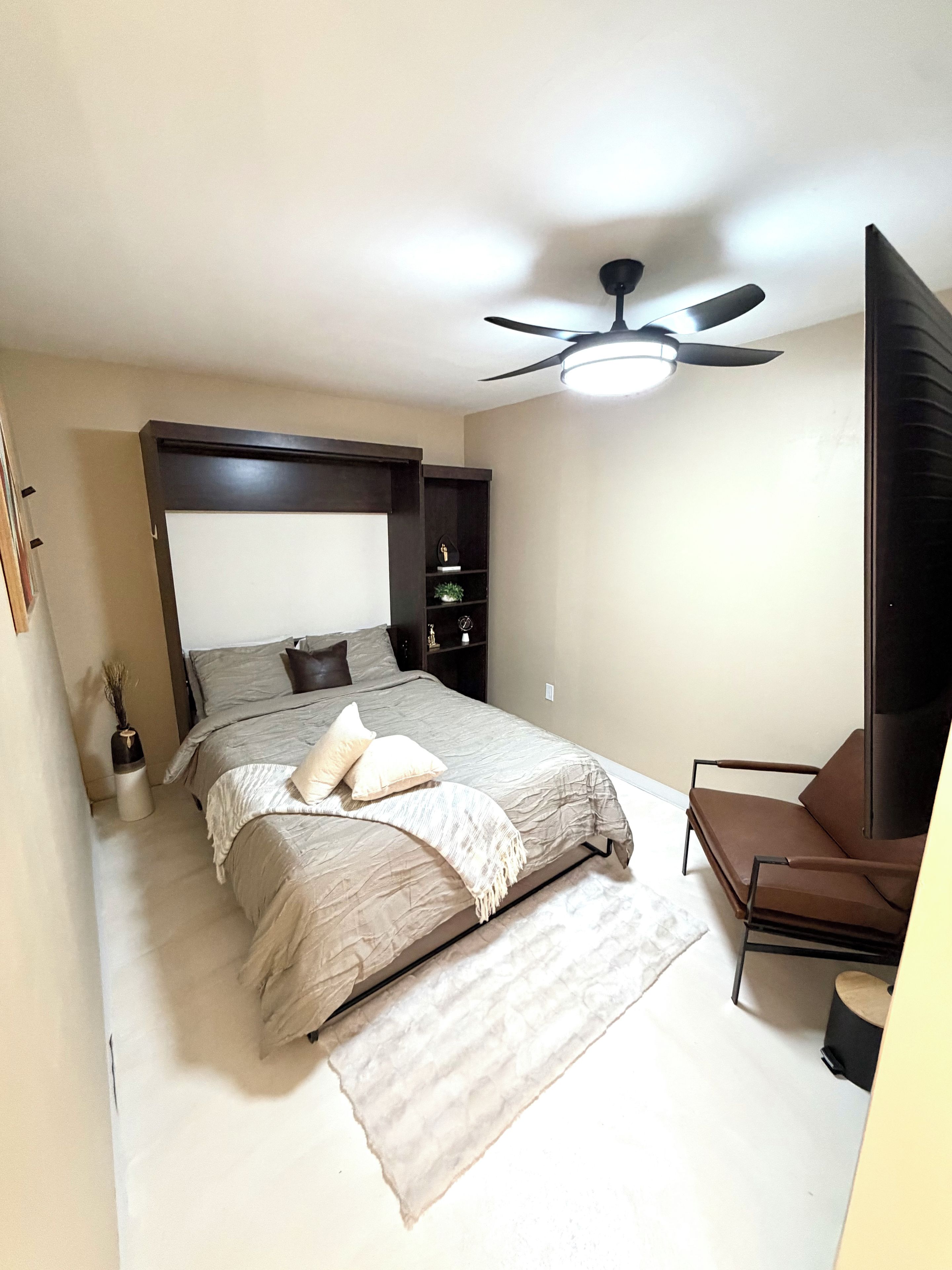 Una camera da letto con un letto, una sedia, una ventola a soffitto e un comodino con una lampada.