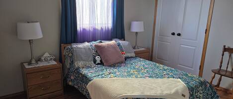 Un dormitorio con una cama, dos mesitas de noche, un aparador, una puerta y una ventana con cortinas.