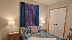 A bedroom with a floral bedspread, two bedside tables with lamps, a wooden nightstand, a door, and a chair. - Pet friendly 2 bedroom and full bathroom with soaker tub close to Westerner. (Red Deer)