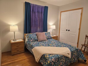 A bedroom with a floral bedspread, two bedside tables with lamps, a wooden nightstand, a door, and a chair. - Pet friendly 2 bedroom and full bathroom with soaker tub close to Westerner. (Red Deer)
