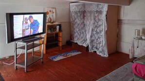 Ein Zimmer mit Fernseher auf einem Metallständer, einem Spitzendesign-Vorhang, einem kleinen Tisch mit Lampe und einem Holzschrank.