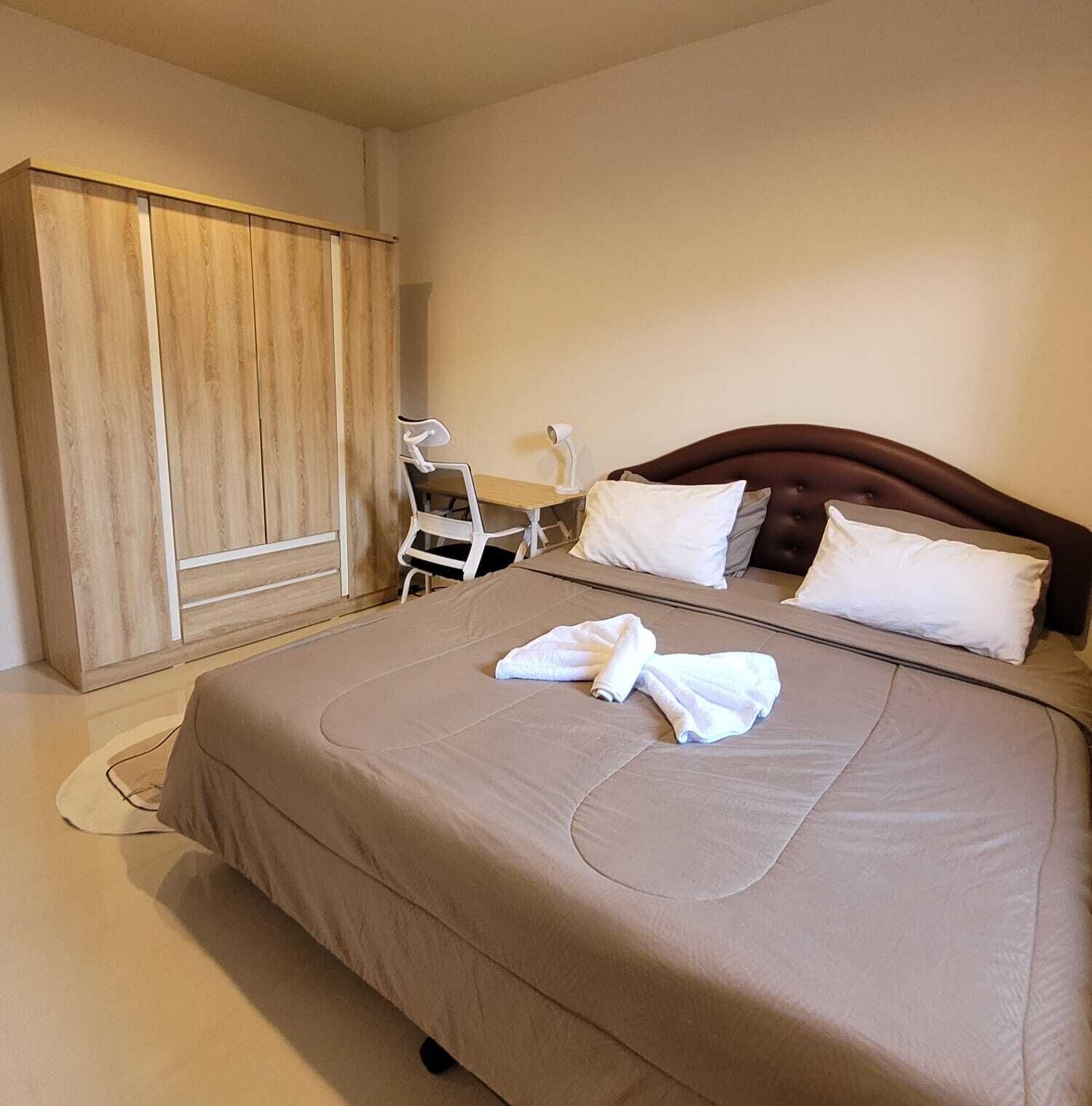 Ein ordentlich bezogenes Bett mit weißen Kissen und einem gefalteten Handtuch, ein Holzschrank und ein weißer Stuhl in einem modernen Hotelzimmer.