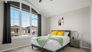 A bedroom with a large window, a bed with a green blanket, a nightstand, and a lamp.