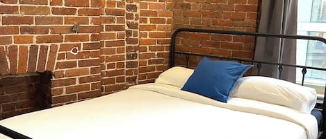 A single bed with a metal frame, white bedding, and a blue pillow in a room with exposed brick walls and a framed picture on the wall.
