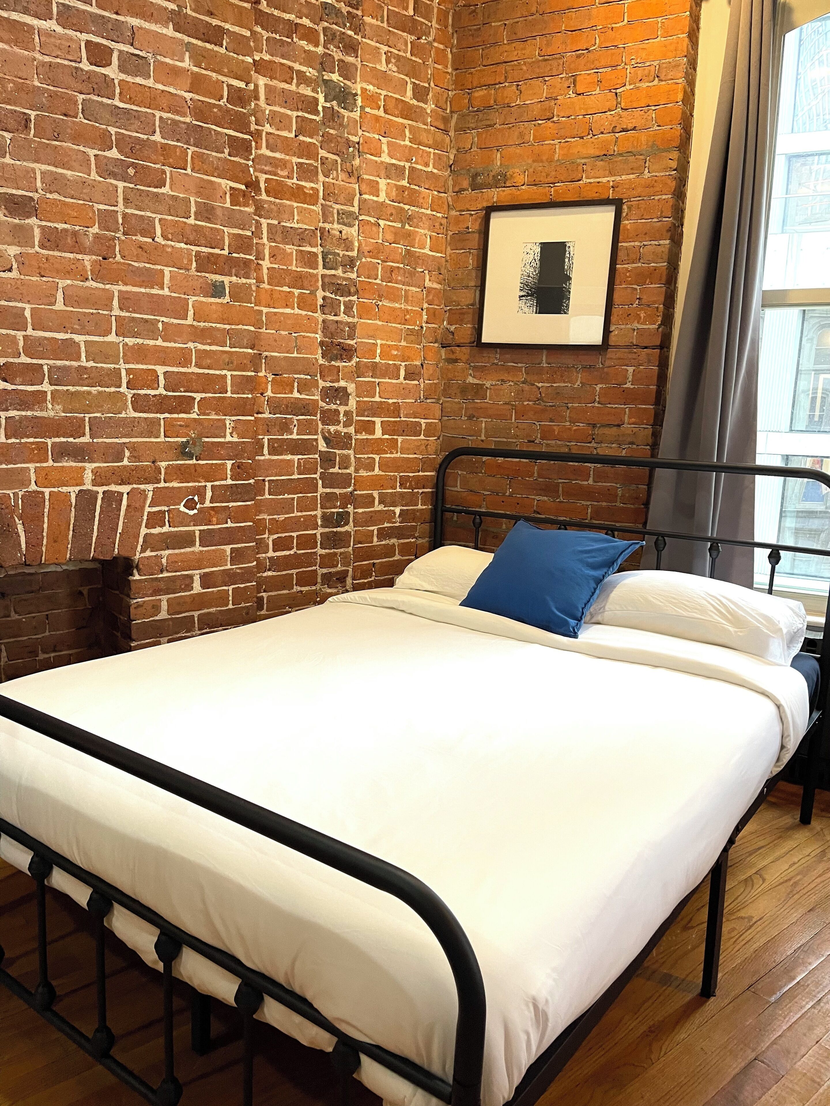 Una cama individual con estructura de metal, ropa blanca de cama y una almohada azul en una habitación con paredes de ladrillo visto y un cuadro en la pared.