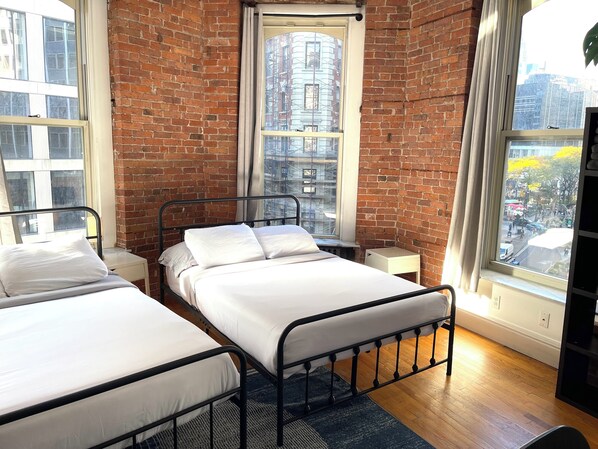 A room with two beds, a brick wall, and large windows with city views. - Nest Aparthotel kitchenette  (New York)