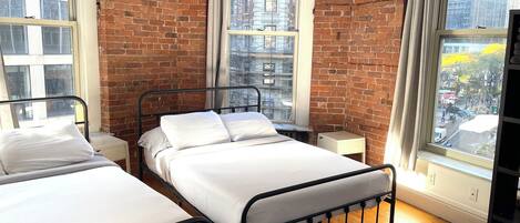 A room with two metal beds, a brick wall, and large windows offering city views.