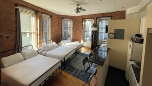 A compact hotel room with two single beds, a small kitchenette, a dining table, and a ceiling fan. - Nest Aparthotel kitchenette  (New York)