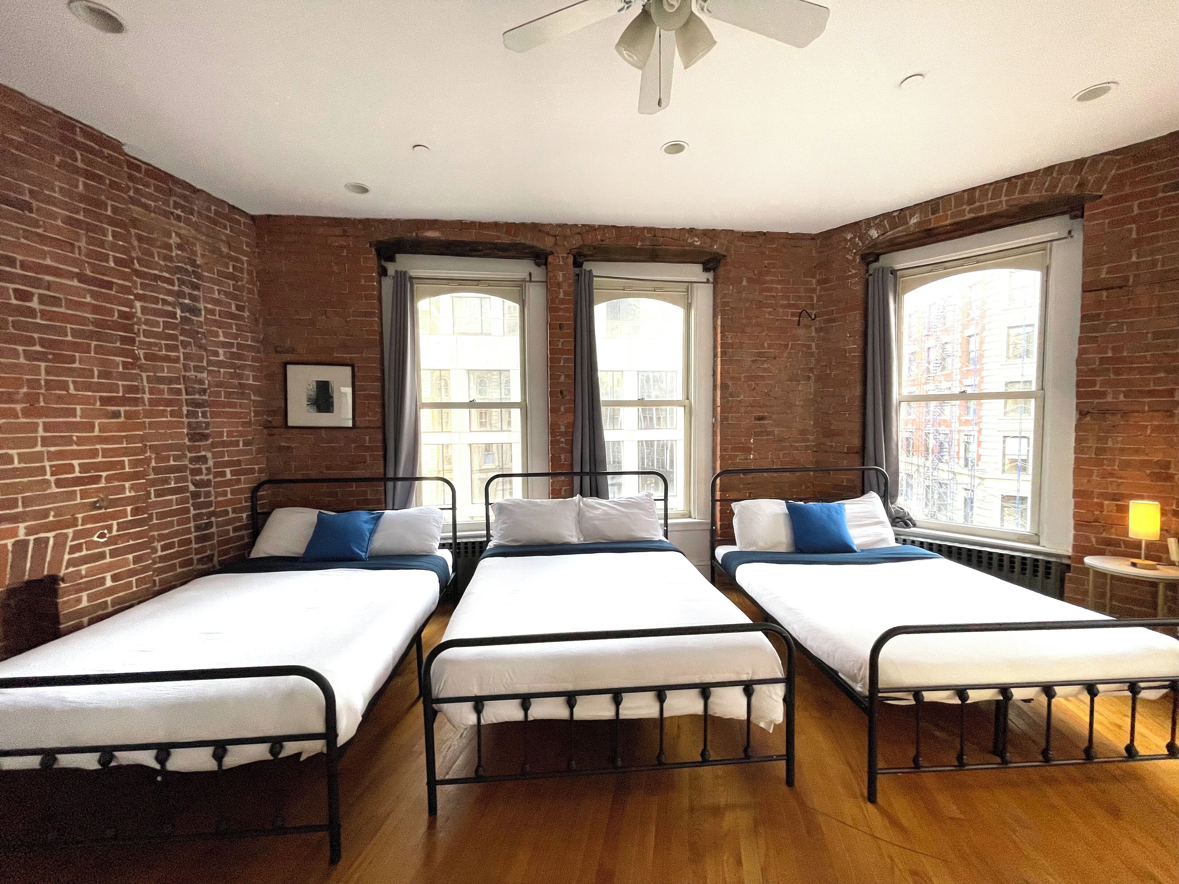A room with three single beds, brick walls, large windows, and a ceiling fan.