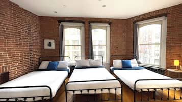 A room with three single beds, brick walls, large windows, and a ceiling fan.