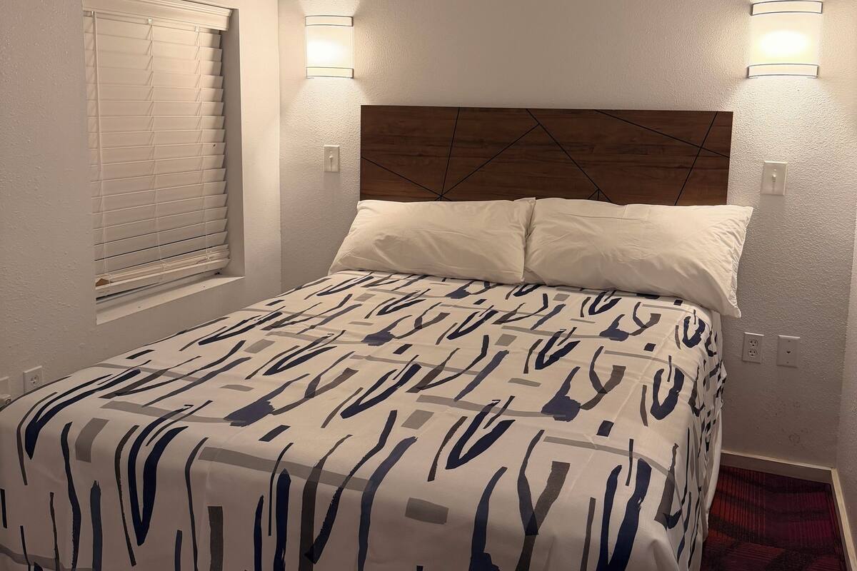 Une chambre d’hôtel avec un lit, deux lampes fixées au mur, une fenêtre avec des stores et un sol recouvert de moquette.