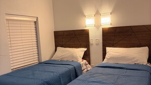 Ein Hotelzimmer mit zwei Betten, jeweils mit blauer gesteppter Bettdecke und weißen Kissen. An der Wand über den Betten befinden sich zwei Wandleuchten.