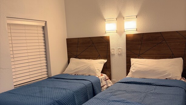 A hotel room with two beds, each with a blue quilted bedspread and white pillows. There are two wall-mounted light fixtures on the wall above the beds.
