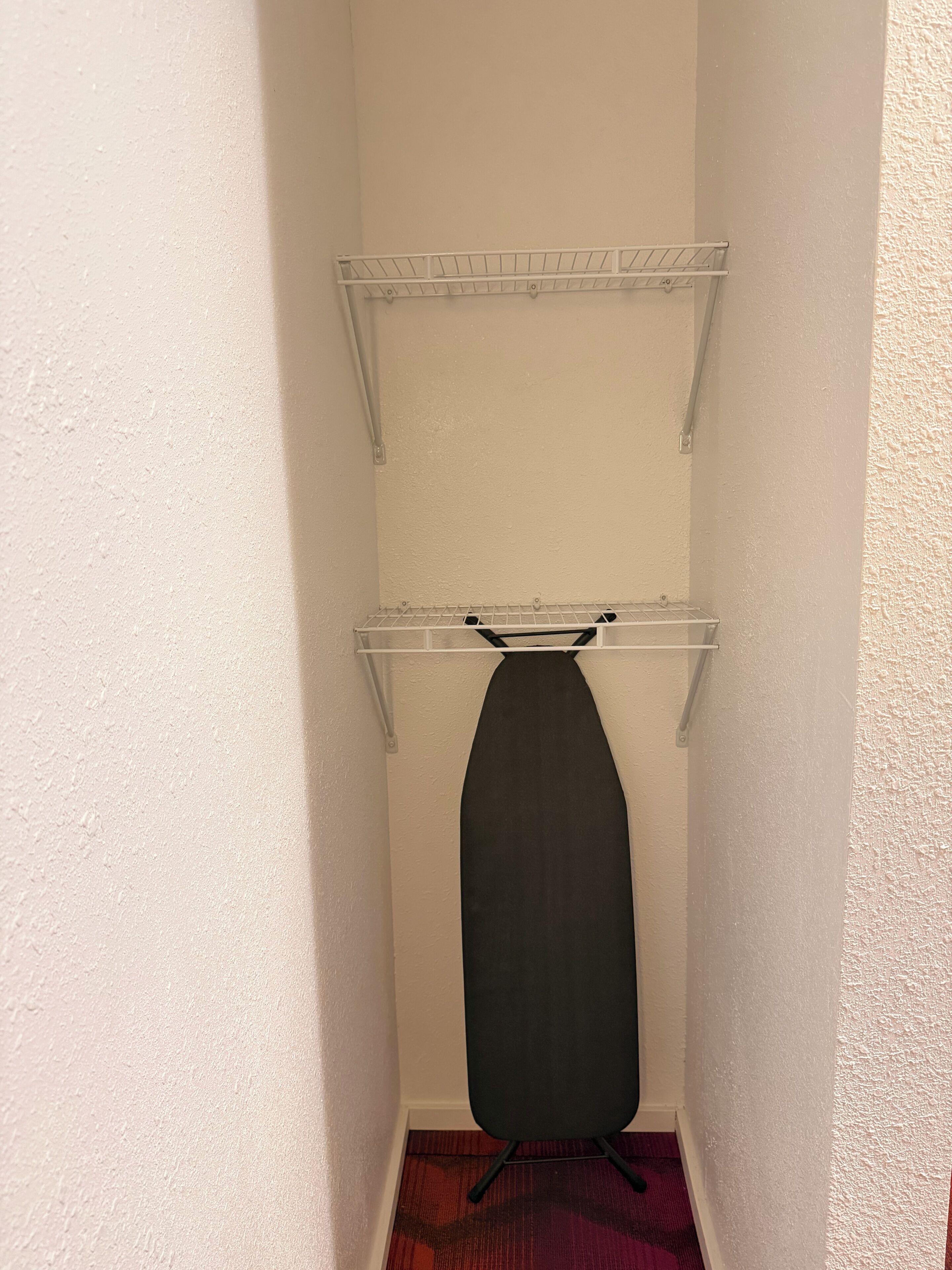 A small room with a black ironing board, two metal shelves, and a red carpet.