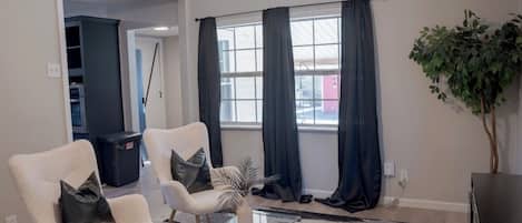Una sala de estar moderna con dos sillones blancos, una mesa de centro de vidrio, un tapete a rayas y un ventanal con cortinas oscuras.